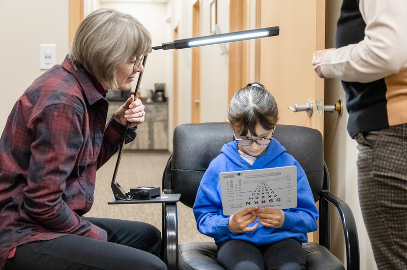 A woman holds a light over a girl reading an eye chart. 