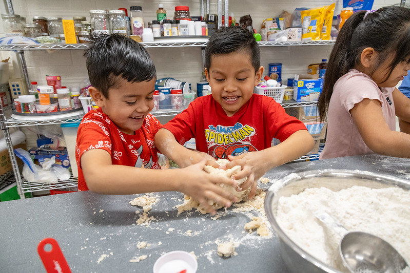 Two young boys smile and laugh while kneading dough together on a table covered in flour. A metal mixing bowl filled with flour and a large spoon sits nearby. In the background, shelves are stocked with baking supplies, ingredients, and kitchen tools.