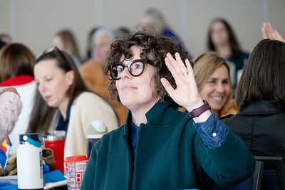 Educators seated at an event, close up of person with glasses and curly hair raising hand