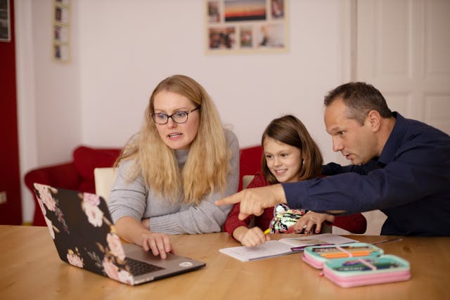 Parents helping daughter in online class, at table with laptop open