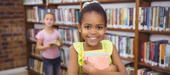 Young girl smiling while holding a book in front of books stacks in library