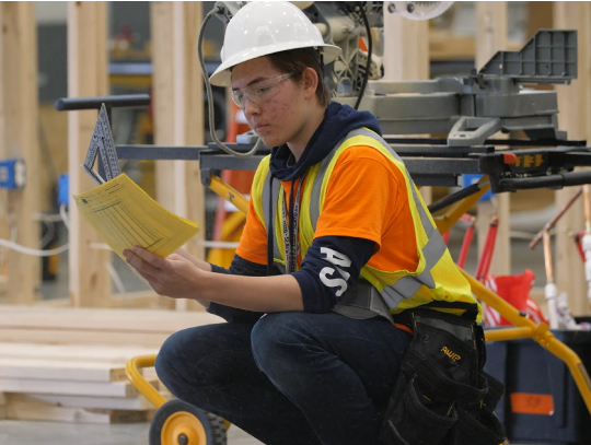 Student in a hard hat reading a manual near heavy equipment