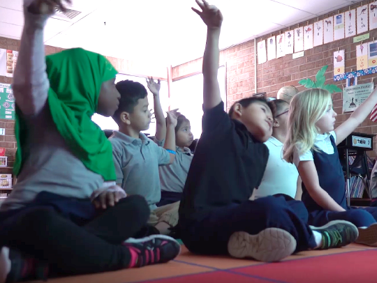Young students sitting on the floor in a classroom and raising their hands to participate