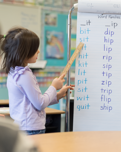 Young girl in classroom pointing to a chart of 3 letter rhyming words with a ruler